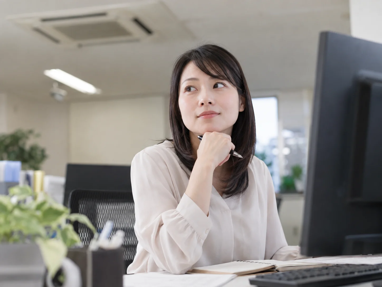 オフィスのデスクで頬杖をつき、ペンを持ちながらモニターから目を離す女性のイメージ