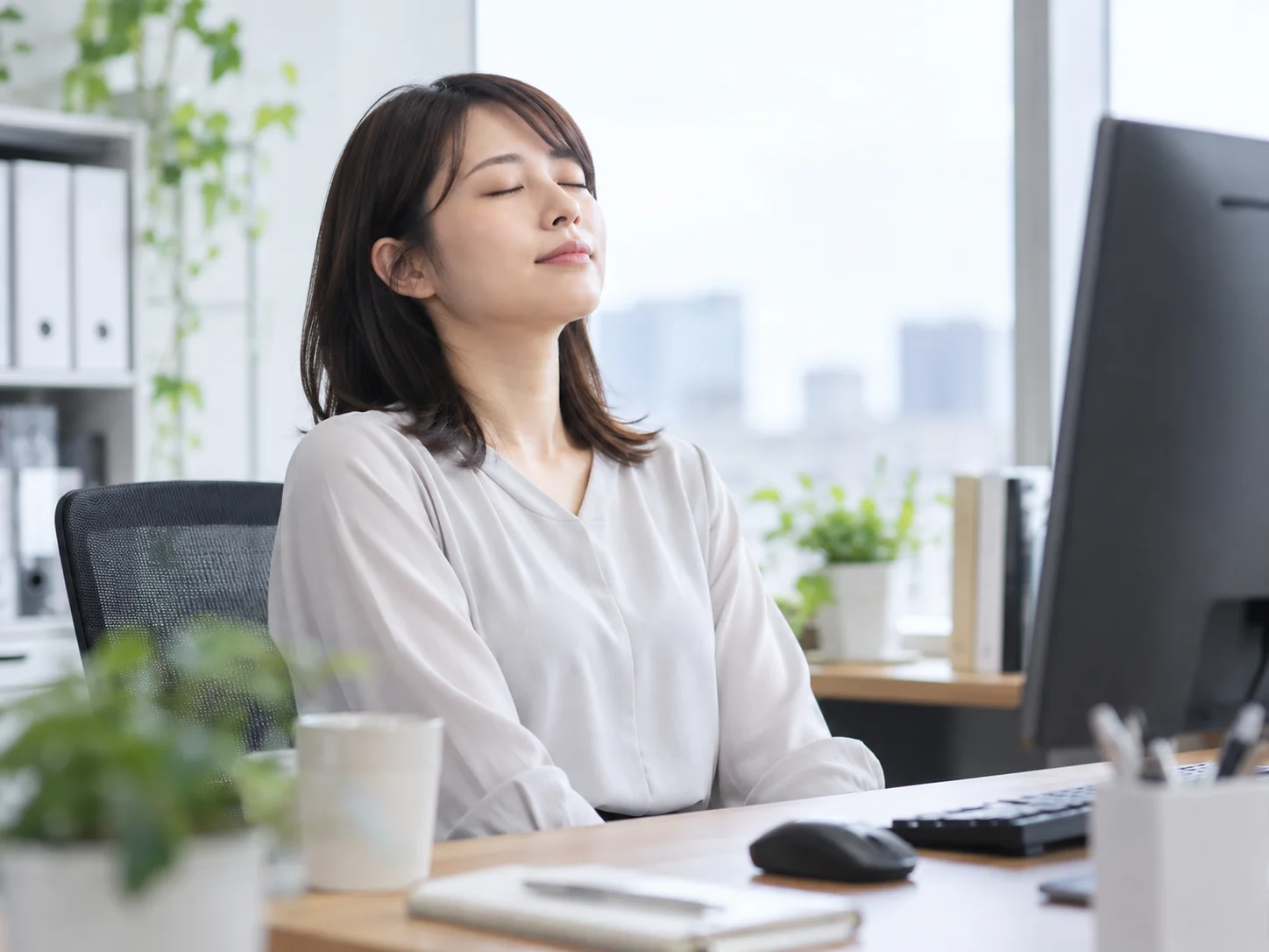 オフィスのデスクでモニターに向かい、目を閉じて上を向く若い女性のイメージ