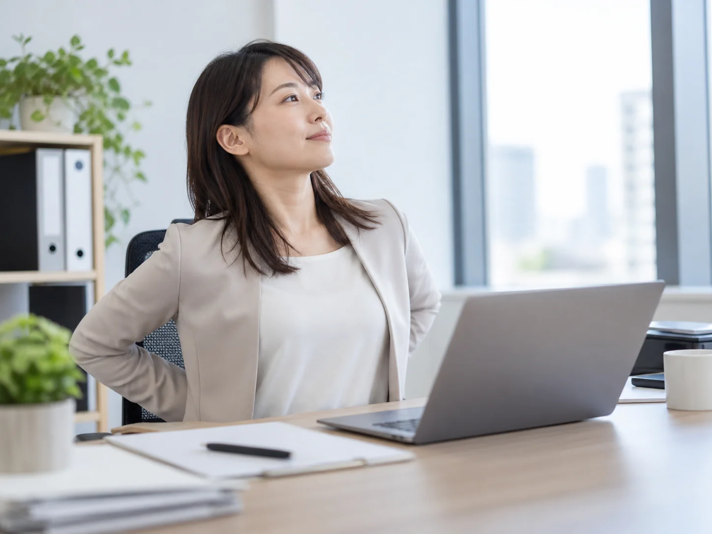 オフィスのデスクで椅子に腰かけ、背中に手を回して伸びをする女性のイメージ