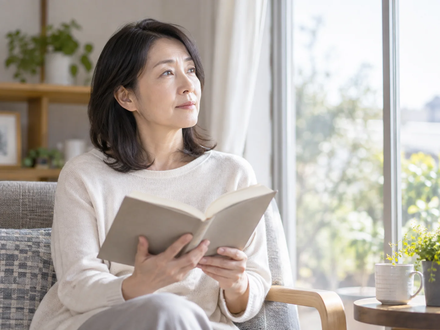 リビングのソファで本を手に持ち、視線を上げて窓の外を眺める中年の女性のイメージ