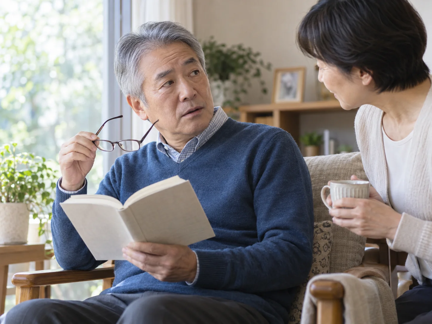 明るいリビングの椅子で隣の女性と話す、本と眼鏡を手にした高齢の男性のイメージ