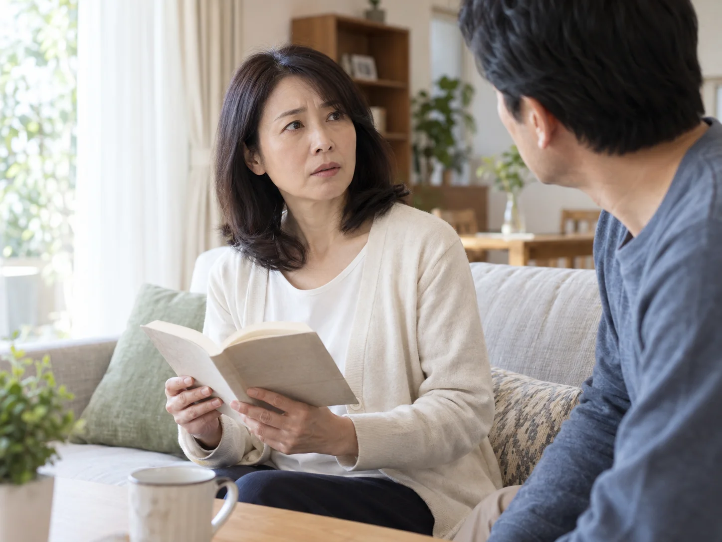 リビングのソファで本を手にして向かいに座る男性と向き合う女性のイメージ